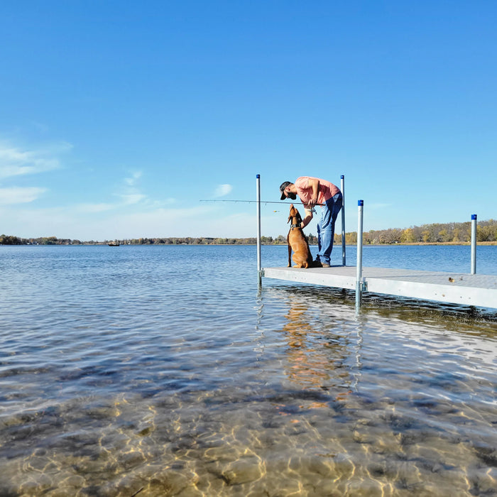 Man and dog on a dock by a lake with clear water and blue sky.